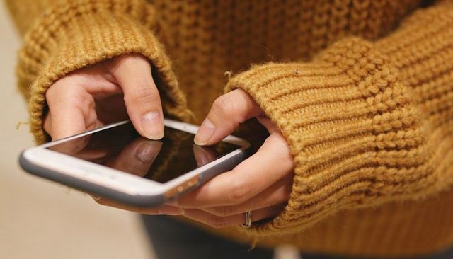 Tapping smartphone screen with cozy mustard knit sweater and silver ring close-up