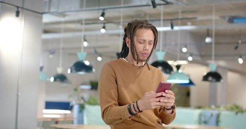 African American man checking smartphone in modern coworking lounge wearing brown sweater