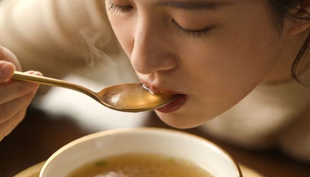 Close-up asian woman sipping steaming soup with gold spoon, cozy home dining moment