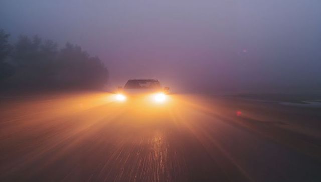 Car approaching through dense fog with warm headlights reflecting on rain-slick rural road