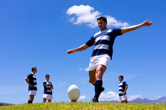Caucasian male rugby player on a pitch during a rugby game on a sunny day, kicking a ball, his teammates in the background. Sports athletic competition.
