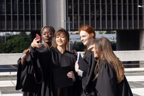 Diverse female graduates taking selfie on urban campus after commencement ceremony