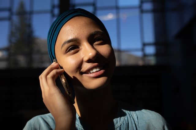 Young biracial female student wearing a dark blue hijab is talking on the phone in a library. She is smiling and appears to be enjoying the conversation. This image can be used for educational websites, communication technology promotions, multicultural and diversity campaigns, or student life articles.