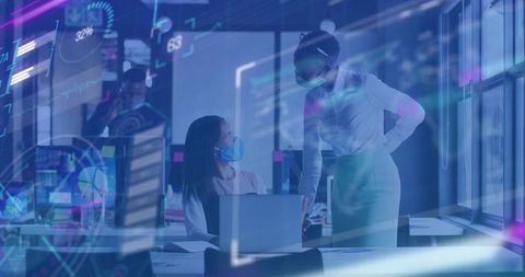 Businesswomen collaborating over laptop in modern masked open-office with data overlays