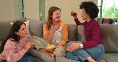 Multicultural women chatting and laughing on cozy couch while sharing snacks and drinks