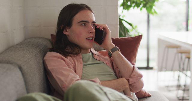 Young person sitting comfortably on a couch, engaged in a phone call. Ideal for use in lifestyle blogs, communication services, phone company advertisements, and casual home-related content.