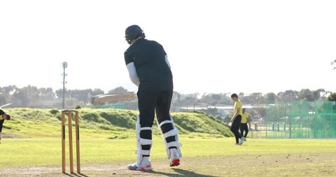 Batsman practicing cricket on sunny field wearing helmet, pads and wooden bat
