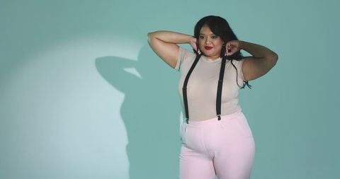 Curvy woman posing wearing black suspenders and pale pink trousers on teal studio backdrop