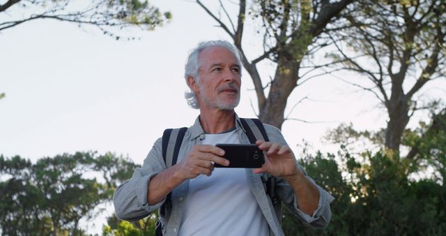 Caucasian man captures memories with his smartphone, outdoor. He enjoys a sunny day of adventure, immersed in nature's beauty.