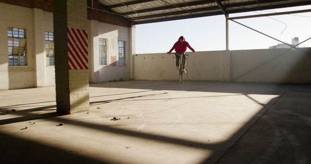 This depicts a man performing a stunt on a BMX bicycle inside an abandoned warehouse. Sunlight streams through the high windows, casting stark shadows on the concrete floor. Suitable for use in articles, advertisements, and websites related to extreme sports, skateparks, urban exploration, or adventure lifestyles.