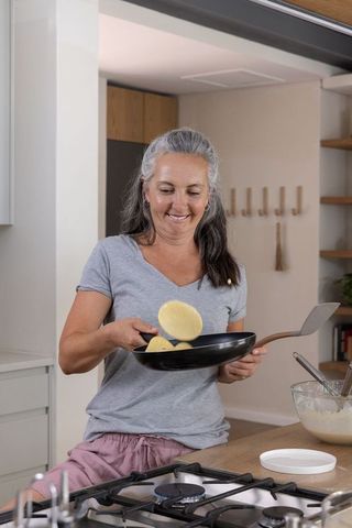Mature woman flipping pancakes while smiling in modern kitchen, casual breakfast moment