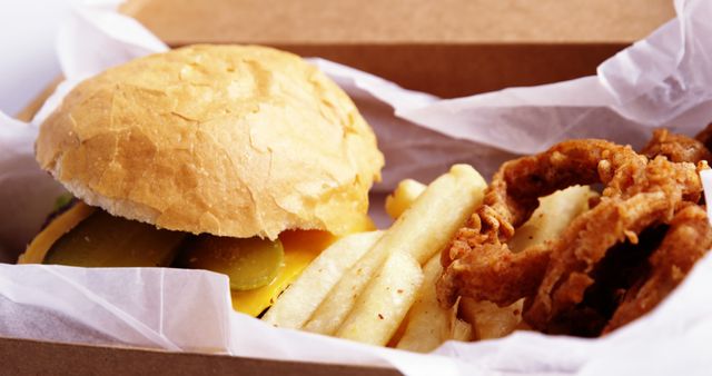 Visual captures enticing cheeseburger with melted cheese and pickles accompanied by golden onion rings and crispy french fries in a paper-lined takeout box. Ideal for marketing restaurant menus, food delivery services, fast-food advertisements, and promotions for casual dining experiences.