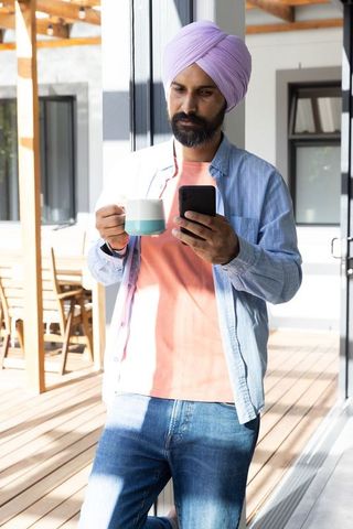 Mid adult man with turban enjoying coffee and smartphone outdoors on deck
