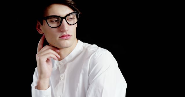 Depiction of young man wearing eyeglasses and a white casual shirt against black background, appearing contemplative and serious. This can be used in fashion blogs, eyewear advertisements, or articles focused on male fashion or contemplation.