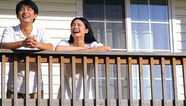 Laughing couple leaning on wooden balcony railing, sunlit home exterior lifestyle moment