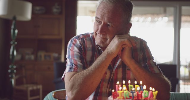 Senior man in plaid shirt seated indoors, reflecting during his birthday celebration with a cake in the foreground. This touching and introspective scene shows the deeper thoughts accompanying a milestone day. Ideal for use in themes of aging, birthdays, family moments, and life reflection.
