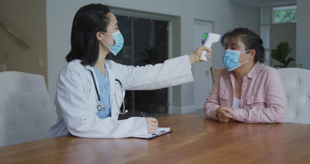 Doctor checking patient's temperature using infrared thermometer, both wearing masks indoors. Suitable for healthcare, medical examination, clinic, COVID-19 protocols, protective gear, patient care, professional consultation.