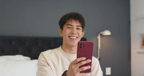 Asian man smiling while holding maroon smartphone on bed, relaxed casual home lifestyle
