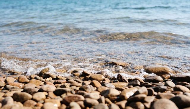Sunlit pebble shoreline with clear shallow water, gentle ripples and foamy edge