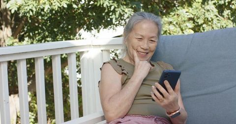 Senior asian woman smiling while checking smartphone on sunny porch