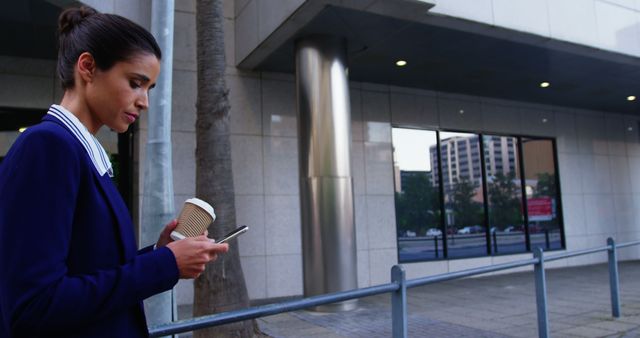 This visual represents a businesswoman holding a coffee cup and using a smartphone while standing outside in an urban area. It's perfect for illustrating themes such as work-life balance, morning routines, modern communication, and urban professional life. Use it for corporate materials, tech promotions, lifestyle blogs, and articles about city living.