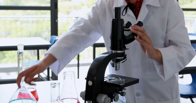 Scientist in a laboratory operating a microscope, with various scientific glassware on the table. Ideal for use in educational materials, scientific presentations, or promotional content for research facilities.