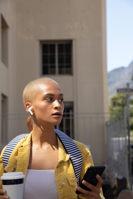Biracial woman with short blonde hair wearing a yellow jacket and striped backpack, using a smartphone with wireless earphones on a sunny day in the city. Ideal for themes of modern lifestyle, urban living, independence, and technology use.
