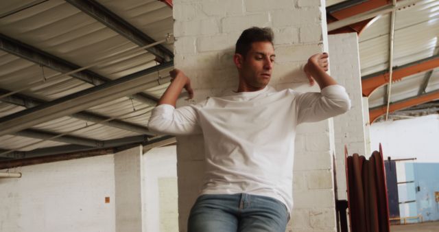 Man in casual attire, relaxing against white brick wall in a warehouse. This image can be used for fashion, lifestyle, or urban-themed projects. Suitable for advertising casual clothing brands, promoting urban living, or use in articles about modern industrial spaces.