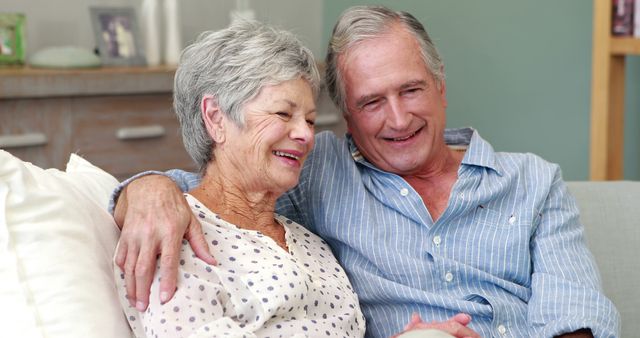 Elderly couple sitting on couch, embracing and smiling while enjoying each other's company. Natural light and homey atmosphere suggest warm, loving relationship perfect for family advertising, healthcare, senior living promotions, or lifestyle blogs.