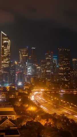 Vertical time-lapsing riverside traffic with light trails and glowing modern skyline
