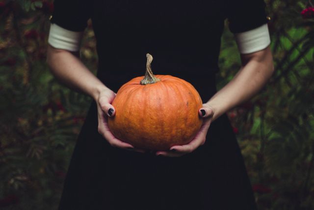 Woman in black dress holding orange pumpkin with both hands in a garden. Green foliage in the background. Ideal for autumn and Halloween themes, gardening blogs, harvest festivals, and seasonal greeting cards.