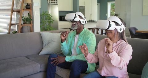 Mature diverse couple wearing VR headsets laughing and exploring virtual reality from couch