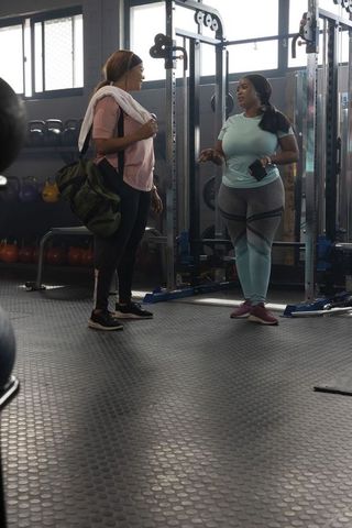 African american women chatting beside power rack in gym with duffel bag and smartphone