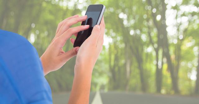 Close-up of hands using a smartphone in an outdoor setting with a blurred forest background. Useful for illustrating connectivity in natural environments, mobile technology usage, and outdoor communication. Could be used for technology advertisements, articles on mobile solutions in rural areas, or lifestyle content regarding combining nature and technology.