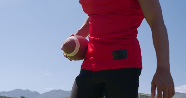 Midsection of biracial male american footballer holding football against sunny blue sky, copy space. Sport, team sport, sports equipment and competition, unaltered.