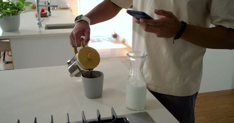 Man pouring coffee into mug while checking smartphone at sunlit modern kitchen island