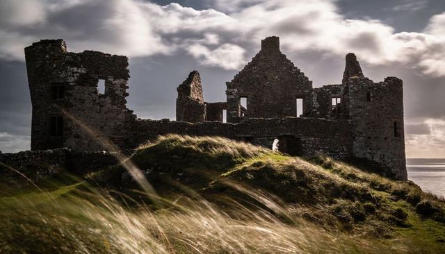 Wind-swept medieval castle ruins on coastal dune with tower silhouettes and moody sky over sea