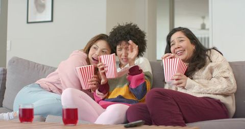 Diverse women laughing and sharing popcorn while watching movie in cozy sweaters at home