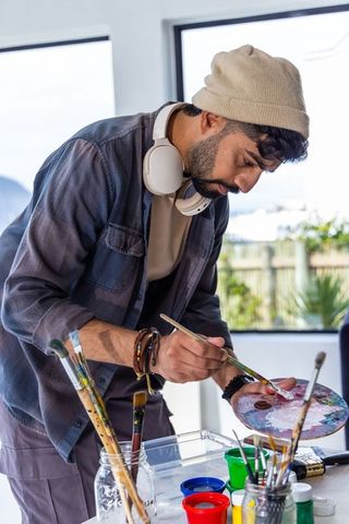 Artist mixing paint on palette while leaning over sunlit studio workspace wearing headphones