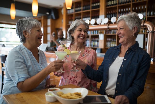 Three senior women are toasting with cocktails in a bar, smiling and enjoying each other's company. This image can be used to depict themes of friendship, socializing, and leisure among older adults. It is suitable for advertisements, articles, or promotions related to senior living, social events, or lifestyle content.
