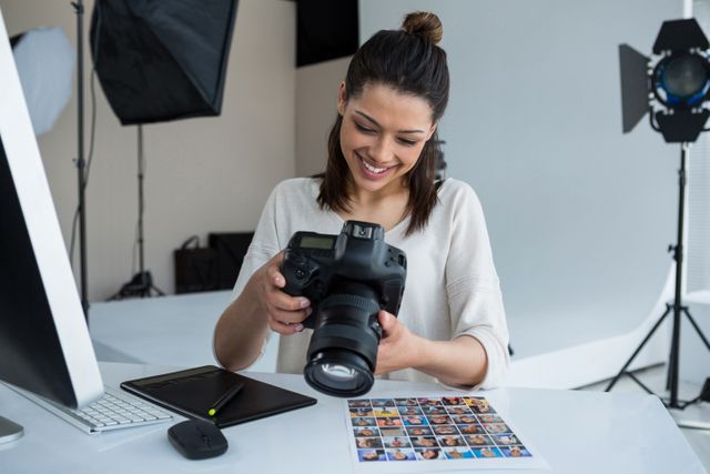 Female photographer is reviewing captured photos on her digital camera at a professional photography studio. The workspace is equipped with a computer, graphics tablet, and printed photo samples. Suitable for illustrating themes related to photography, creative careers, professional work environments, and digital photo editing.
