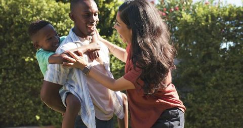 Sunlit backyard family joy playing: father carrying laughing son while partner embraces
