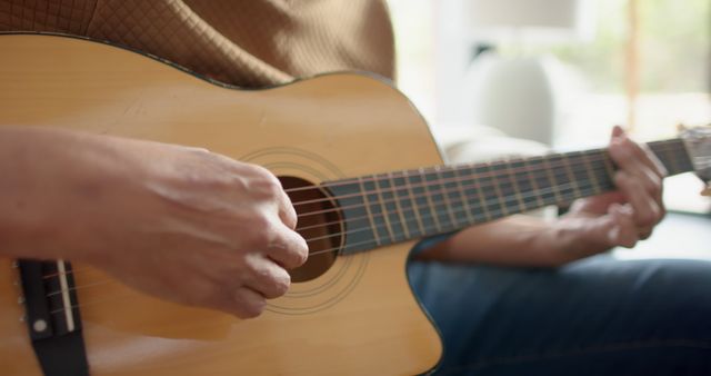 Close-up of hands playing acoustic guitar indoors, highlighting fingers strumming and positioning on strings. Ideal for use in articles related to music, guitar tutorials, hobbies, or as a visual aid for learning materials or advertisements promoting musical instruments.