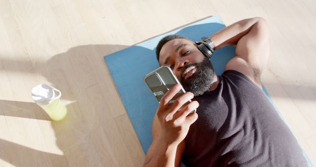Young man in athletic attire relaxing on yoga mat, using smartphone, and wearing wireless headphones. Bright sunlight casts shadows. Next to him, a water bottle indicates hydration. Ideal for themes related to exercise, fitness, technology use during relaxation, personal well-being, or gym settings.