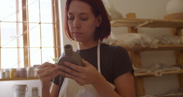 This image shows a young female artisan with red hair deeply focused on crafting a ceramic pot in her studio. Ideal for content related to handmade crafts, artisan work, pottery classes, creative practices, and the art of pottery making.