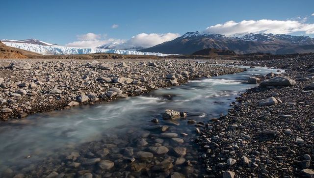 Flowing glacial meltwater carving pebble-strewn riverbed below glacier, snow-capped peaks
