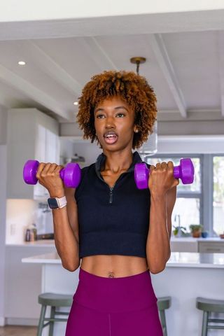 African american woman lifting purple dumbbells in modern kitchen wearing crop top
