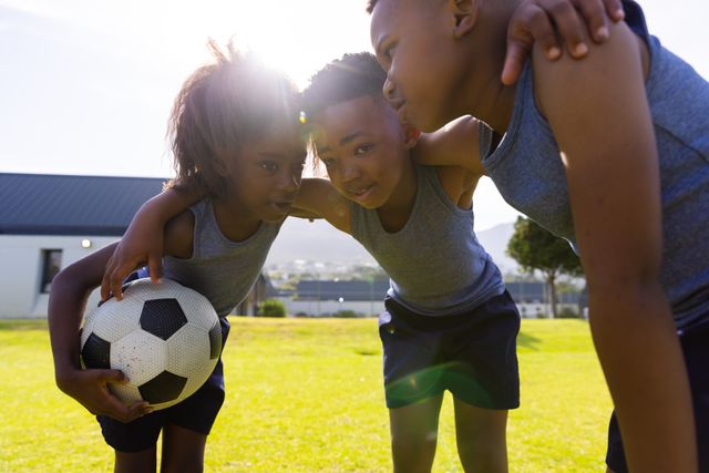 Children huddling with a soccer ball on a school field, showing teamwork and unity. Ideal for educational materials, sports programs, childhood development articles, and advertisements promoting physical activity and teamwork among kids.