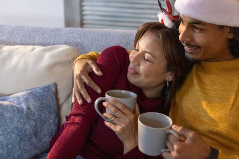 Asian woman and african american man snuggling with hot mugs on couch in holiday sweaters