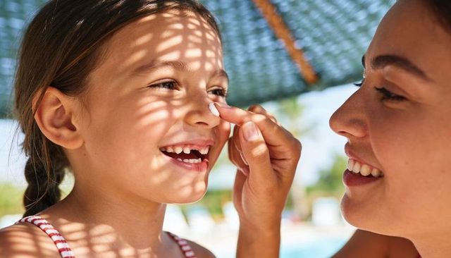 Applying sunscreen to smiling daughter nose under umbrella shadow at poolside summer